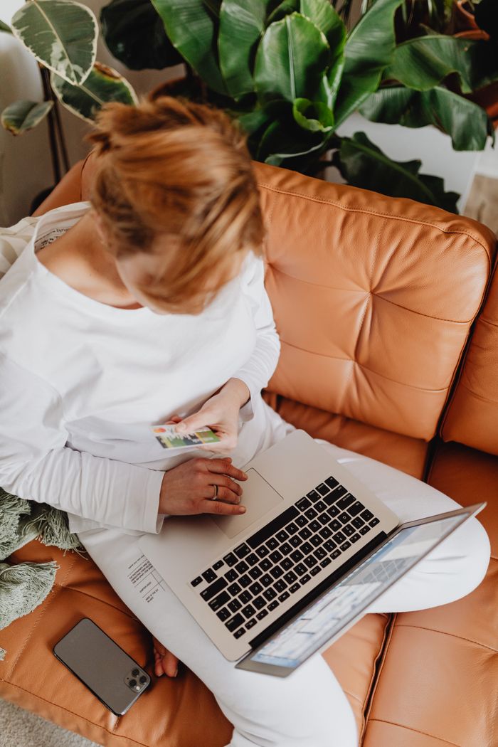 Woman using laptop for online shopping on a cozy sofa surrounded by greenery.