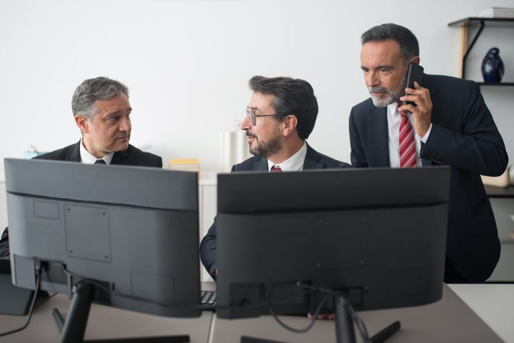 Three businessmen in an office discussing work, two using computers and one on a phone call.